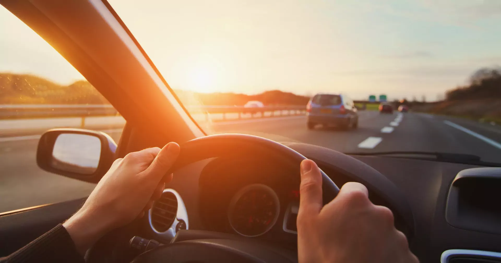 Eine Person fährt bei Sonnenuntergang auf der Autobahn, die Hände am Lenkrad, andere Autos voraus, warmes Sonnenlicht erfüllt den Innenraum des Autos.