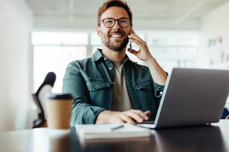 Lächelnder Mann mit Brille benutzt Smartphone und Laptop am Schreibtisch mit Notebook und Kaffee in einem modernen, hellen Büro.