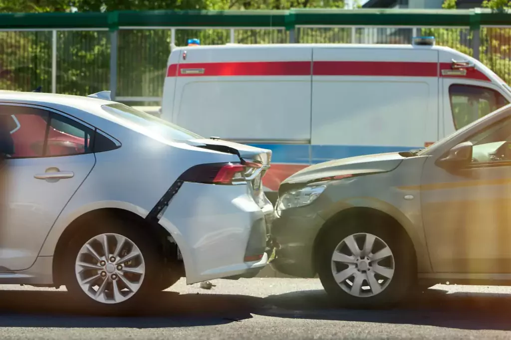 Zwei Autos nach Auffahrunfall mit sichtbarem Schaden; ein silbernes und ein dunkles Auto, Krankenwagen im Hintergrund.