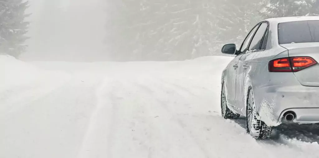 Ein weißes Auto auf einer verschneiten, nebligen und von Bäumen gesäumten Straße; die schlechte Sicht macht die Herausforderungen des winterlichen Fahrens deutlich.