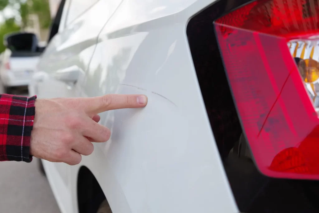 Finger zeigt auf Mietwagenkratzer an der Rückseite eines weißen Autos, nahe dem Rücklicht.