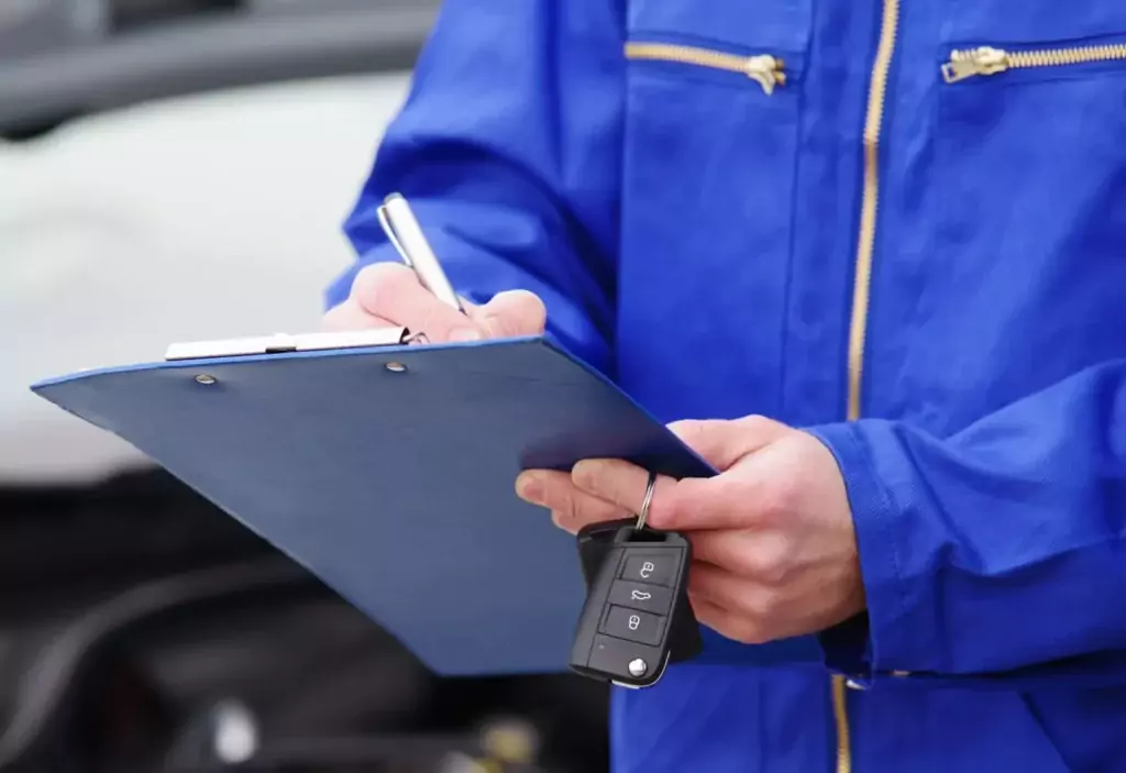 Eine Person in blauer Uniform schreibt auf einem Klemmbrett und hält einen Autoschlüsselanhänger in der Hand; der unscharfe Hintergrund suggeriert eine Fahrzeugkontrolle (TÜV überzogen).