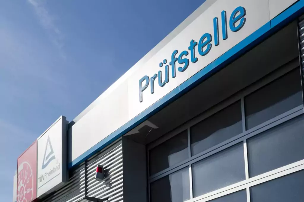 Gebäude mit "Prüfstelle"-Schild, Glasgarage, blauer Himmel, Schilder "TÜV überzogen", Sicherheitsgerät an der Wand.