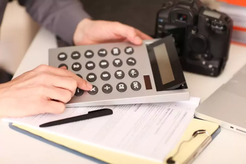 Person drückt Tasten eines großen Taschenrechners auf Bürobedarf, darunter Stift, Digitalkamera und Laptop am Schreibtisch.