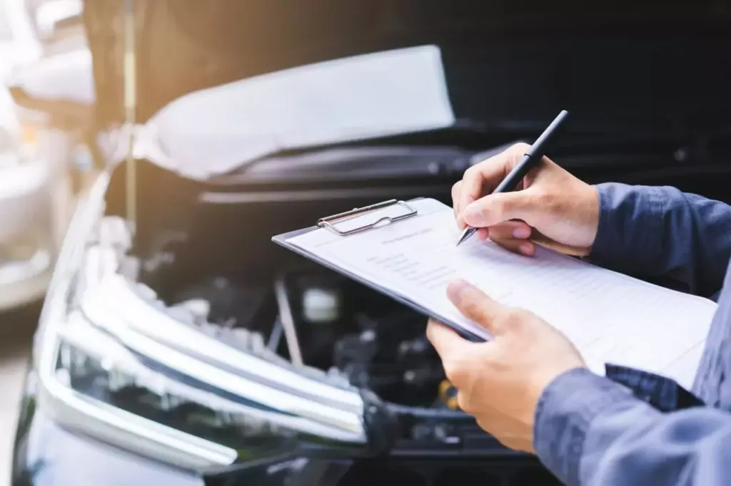 Eine Person mit einem Klemmbrett schreibt ein Formular in der Nähe eines Autos mit offener Motorhaube, das auf eine Fahrzeuginspektion oder -wartung hinweist.