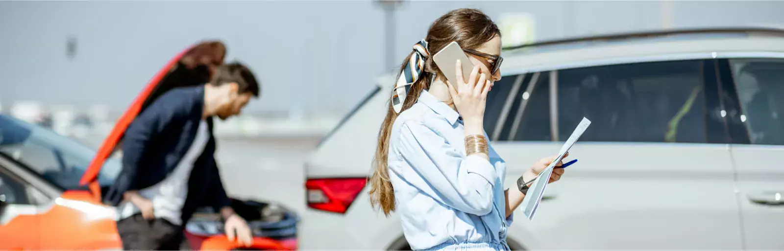 Frau am Auto telefoniert mit Papieren; Mann schaut unter der Motorhaube des roten Autos nach - es scheint sich um einen Notfall am Straßenrand zu handeln.