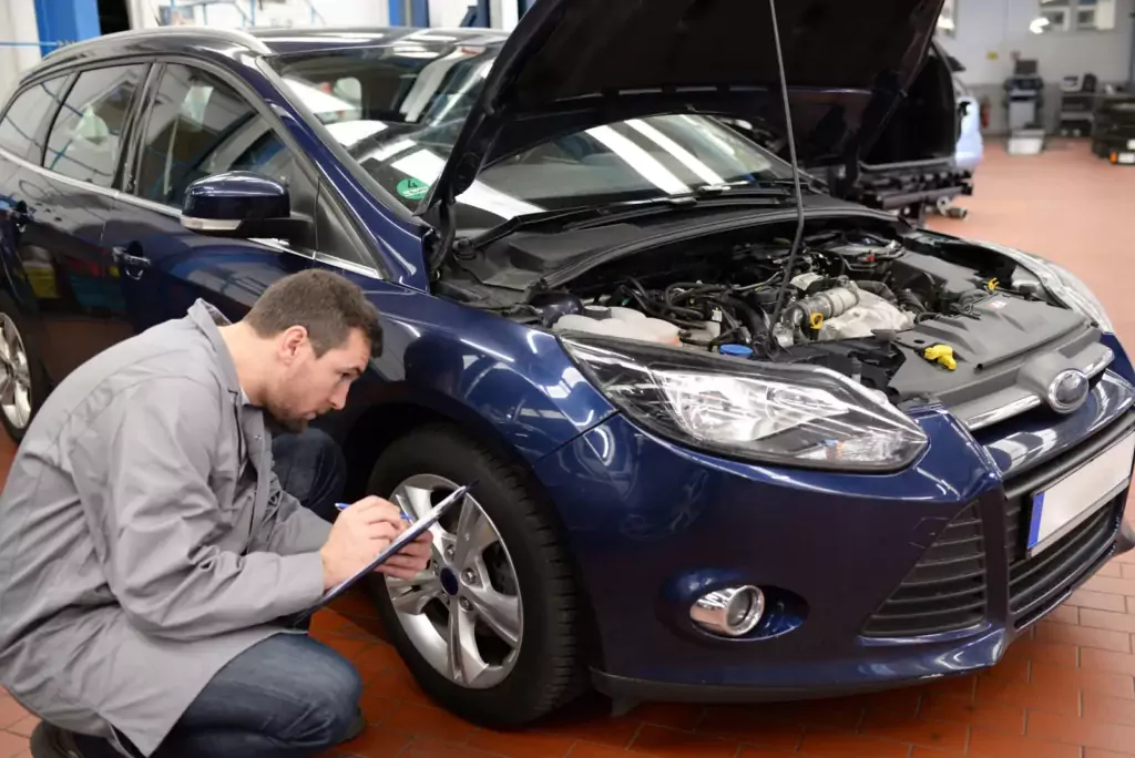 Ein Mechaniker untersucht den Vorderreifen eines blauen Autos und macht sich Notizen auf einem Klemmbrett in der Werkstatt, die Motorhaube ist offen.