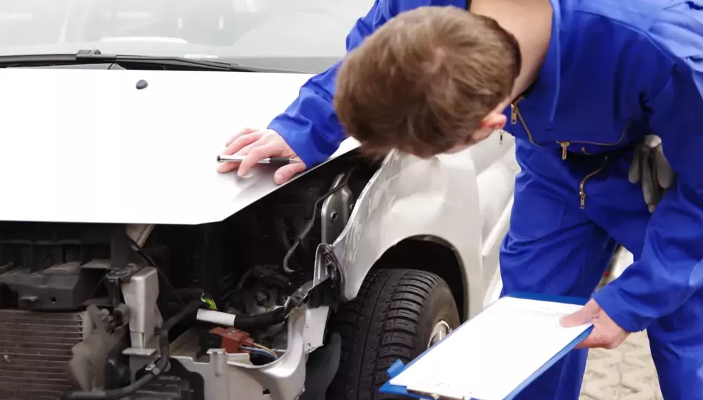 Ein Mechaniker im blauen Overall untersucht den Frontschaden eines weißen Autos mit offener Motorhaube, mit einem Klemmbrett in der Hand, und macht sich Notizen.