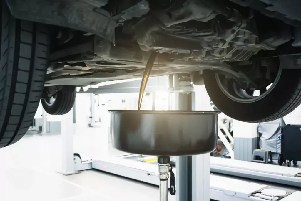 Auto auf Hebebühne, Ölwechsel wird durchgeführt. Motoröl läuft in schwarzen Behältern, helle Werkstattumgebung.
