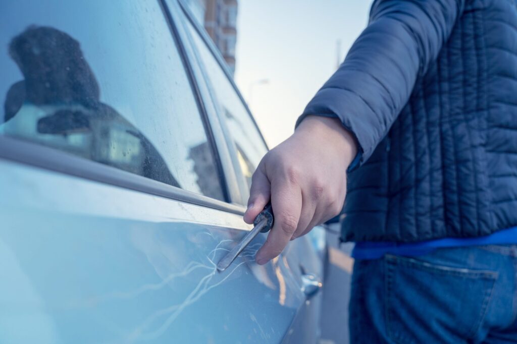 Eine Person in einer Jacke zerkratzt ein blaues Auto mit einem Schlüssel und hinterlässt sichtbare Lackschäden.