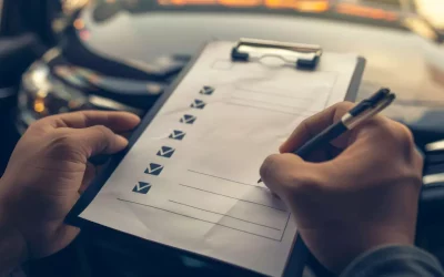 Eine Person schreibt eine Checkliste auf einem Klemmbrett ab; das unscharfe Armaturenbrett eines Autos im Hintergrund deutet auf einen Inspektions- oder Bewertungsprozess hin.
